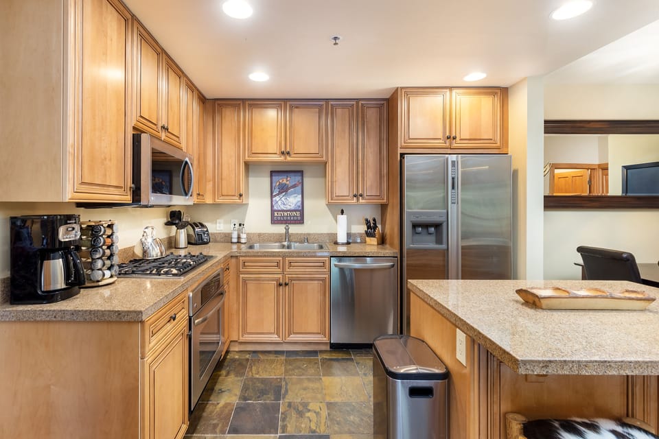 A modern kitchen with light wood cabinets, stainless steel appliances, a black coffee maker, and a kitchen island with a granite countertop. A poster with a wine bottle is on the back wall.