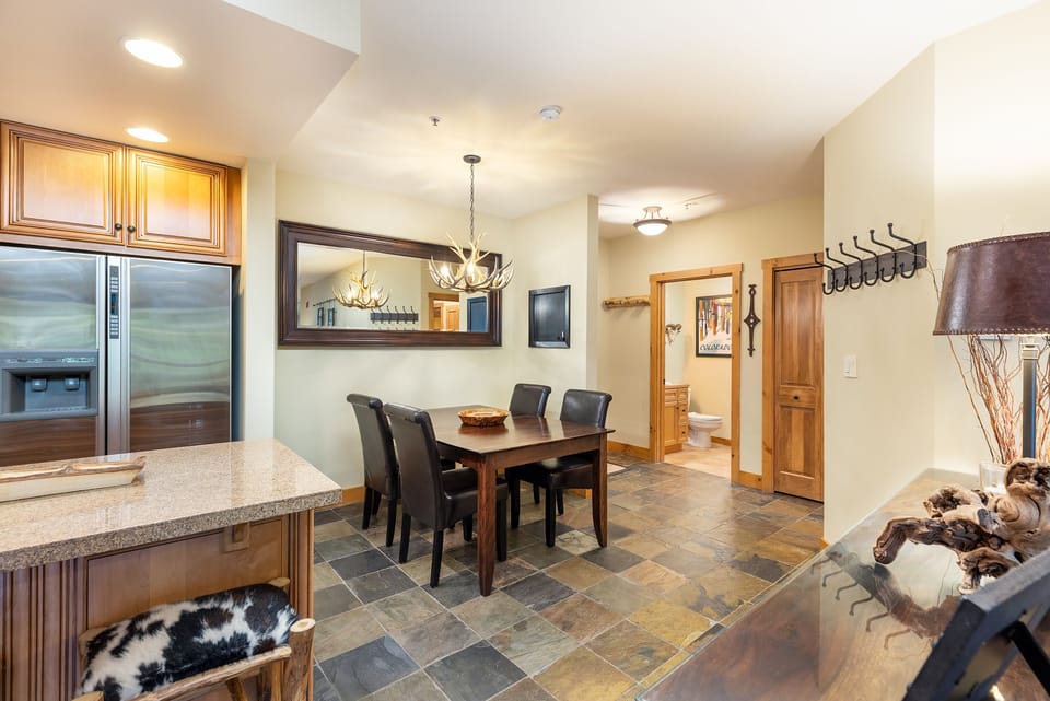 Modern kitchen and dining area featuring wood cabinetry, a large mirror, a rectangular dining table with four chairs, tiled flooring, and a view into a hallway leading to a small bathroom.