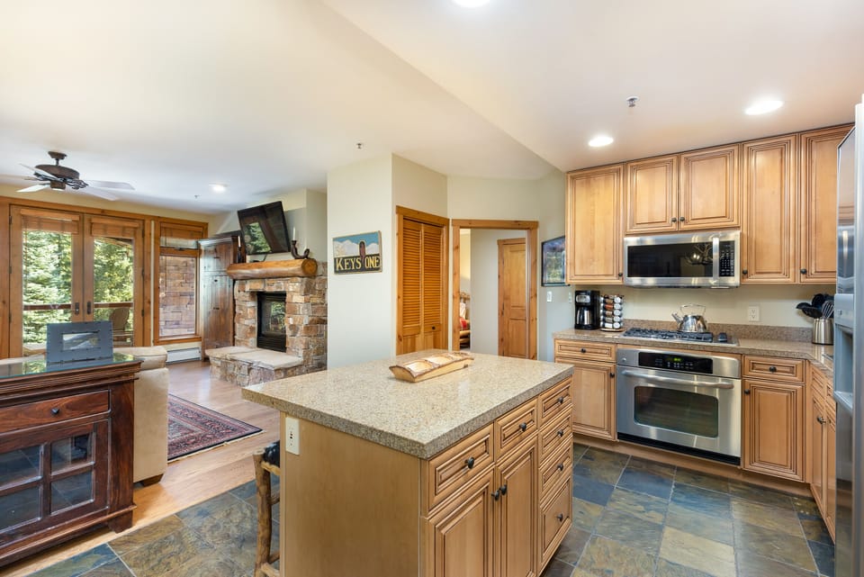 A modern kitchen with wooden cabinets, stainless steel appliances, and a central island. To the left is a cozy living area featuring a stone fireplace, wooden furniture, and large windows.