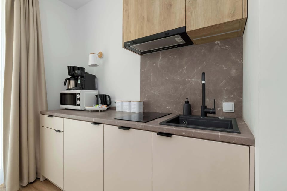 A small kitchenette includes light wood cabinets, a countertop coffee setup, and a dark backsplash. Curtains beside the kitchen area soften the modern design.