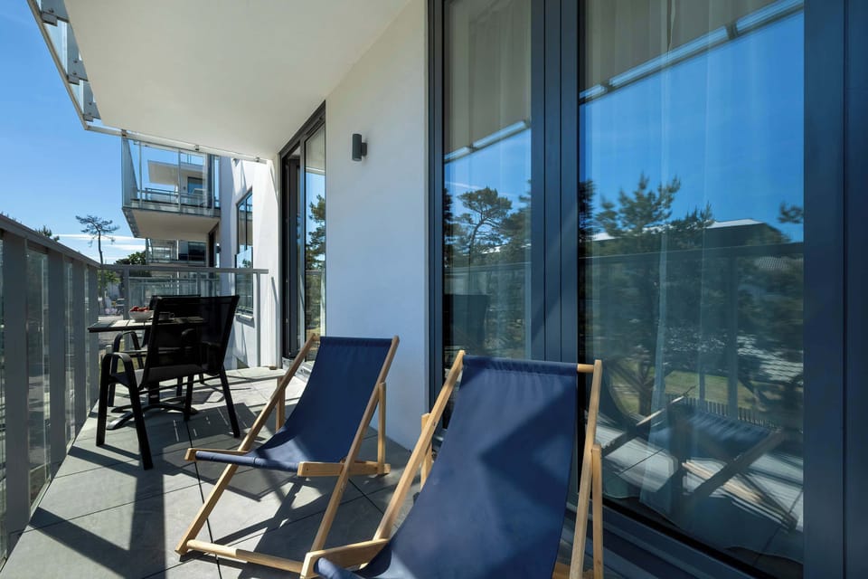 Two blue lounge chairs sit on a balcony facing neighboring buildings and greenery. Glass railings maintain clear views of the surroundings.