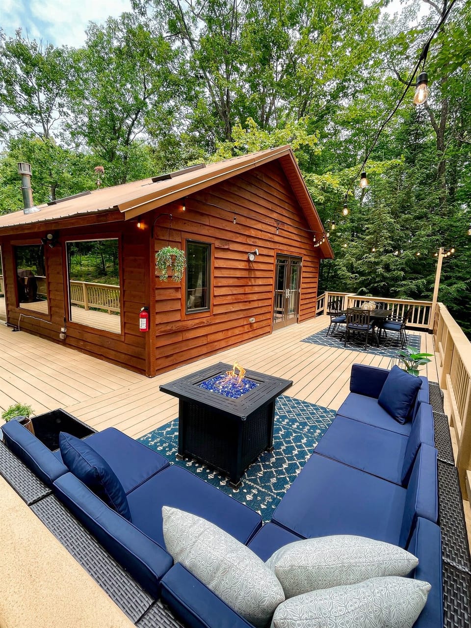 Main cottage back deck feature lounge couch with propane fire pit, 6 person dining table lit by string lights. Wooded views. 