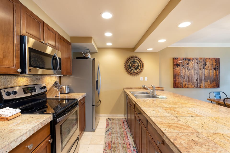 Modern kitchen with stainless steel appliances, wooden cabinets, granite countertops, a center island with a sink, and a wall clock. There is also an art piece on the wall and a dining area.