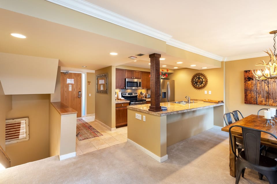 Modern kitchen and dining area with wooden cabinets, stainless steel appliances, granite countertops, and a chandelier over the dining table. The space is open-plan, leading to a hallway and stairs.
