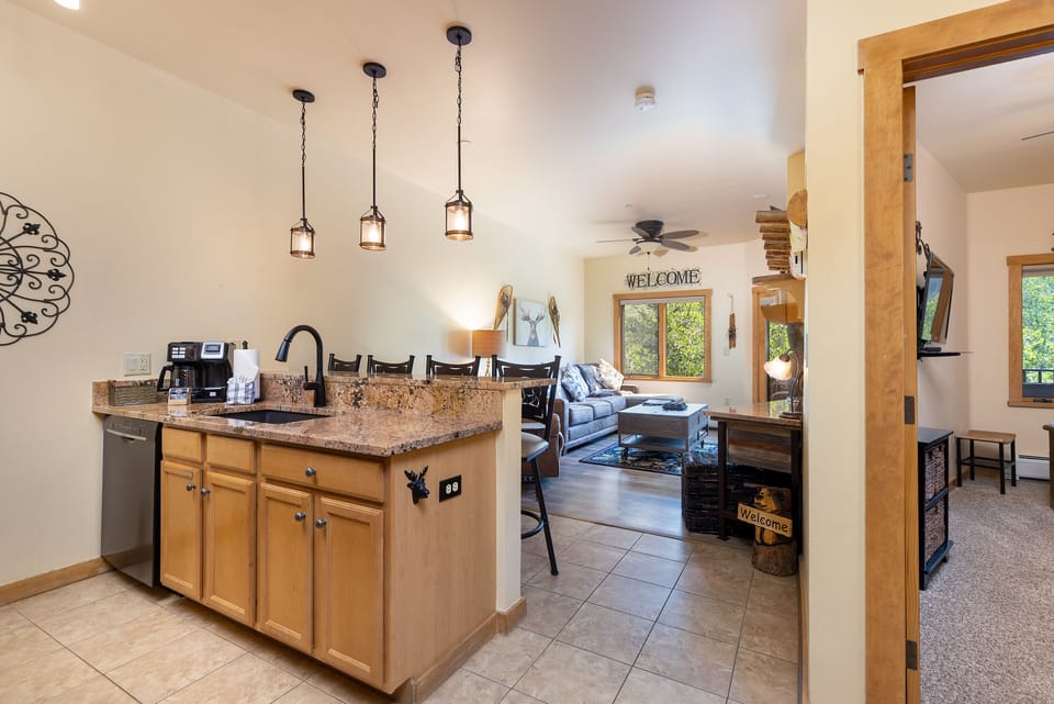 Kitchen and living area of a home featuring a granite countertop island with pendant lights, wooden cabinets, a sofa, and a TV. Large windows provide natural light and there are decorative welcome signs.