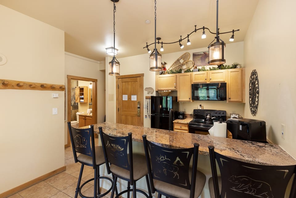 A kitchen with wooden cabinets, black appliances, hanging pendant lights, and a granite countertop with four high chairs. A wooden coat rack is on the left wall and a hallway leads to more rooms in the background.