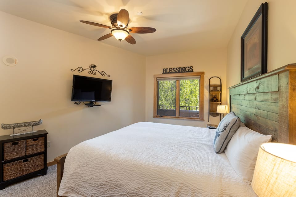 A cozy bedroom features a wooden bed with white bedding, a ceiling fan, a wall-mounted TV, wicker drawers, and a window with a view of trees. The word "Blessings" is displayed above the window.