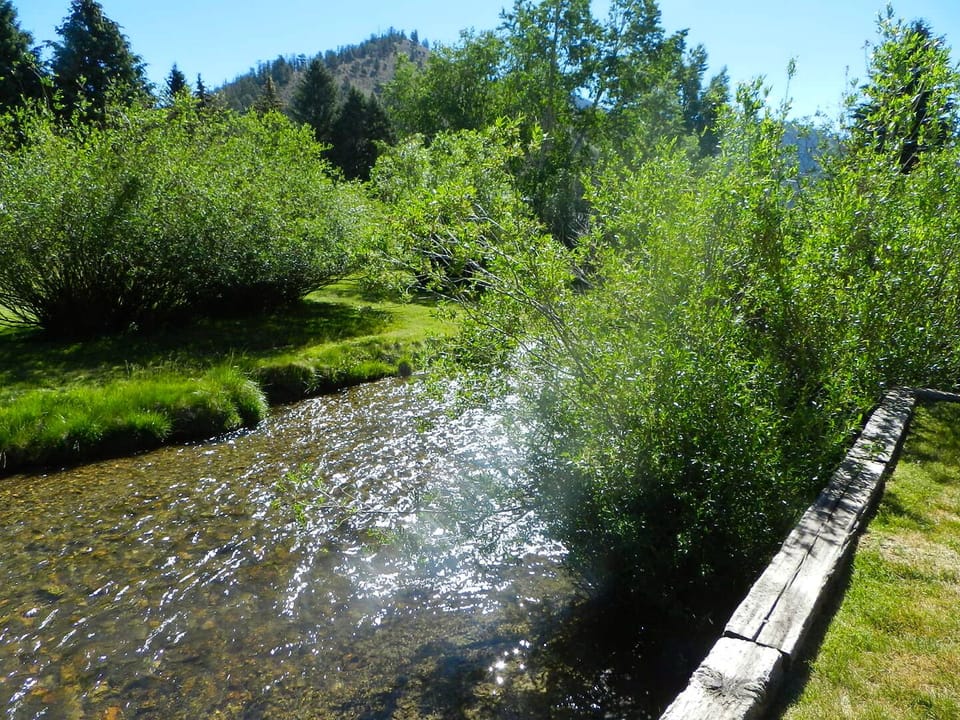 A peaceful creek flows through the property grounds, surrounded by lush greenery and framed by forested hillsides under a clear blue sky.