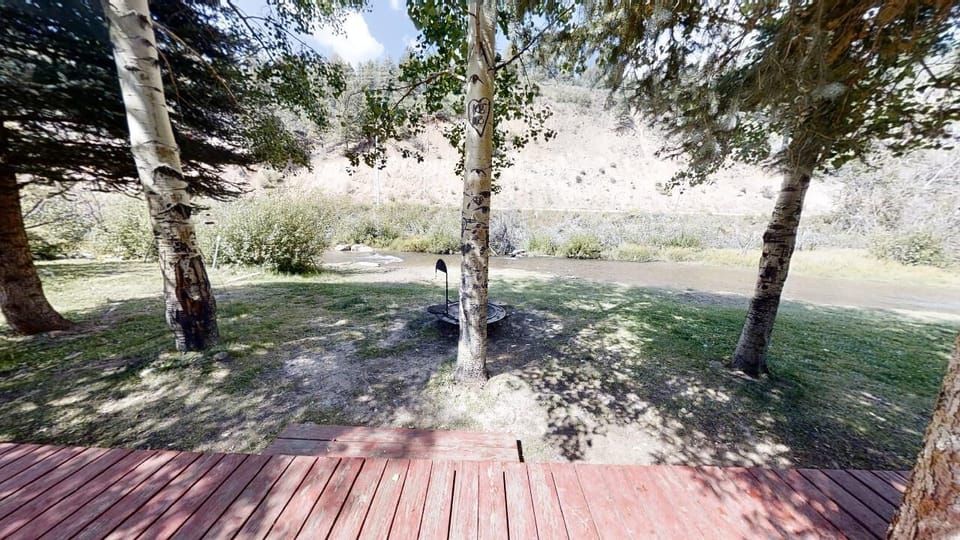 A peaceful riverfront setting with mature birch trees and natural landscape surrounding the property. A wooden deck offers direct access to the serene outdoor space.