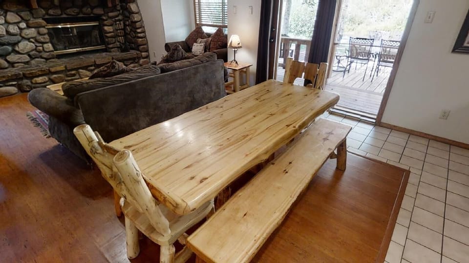 Step into your cozy mountain retreat featuring a rustic log dining table, plush seating by the stone fireplace, and your private deck overlooking natural surroundings—perfect for memorable meals and relaxation.