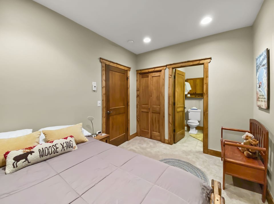 A bedroom with beige walls, a queen-sized bed with pillows and a "Moose X-ing" cushion, dual closets, a doorway to a bathroom with a toilet in view, and a wooden bench with stuffed animals.