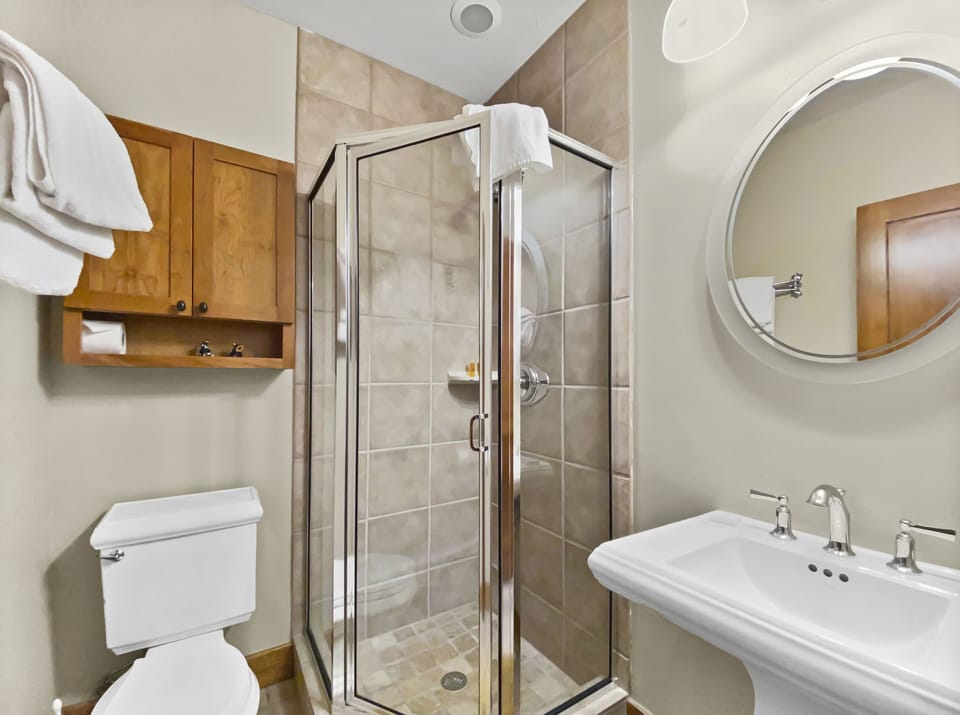 A bathroom with a glass-enclosed shower, a toilet, a pedestal sink, a round mirror above the sink, and a wooden cabinet with towels and toiletry items.