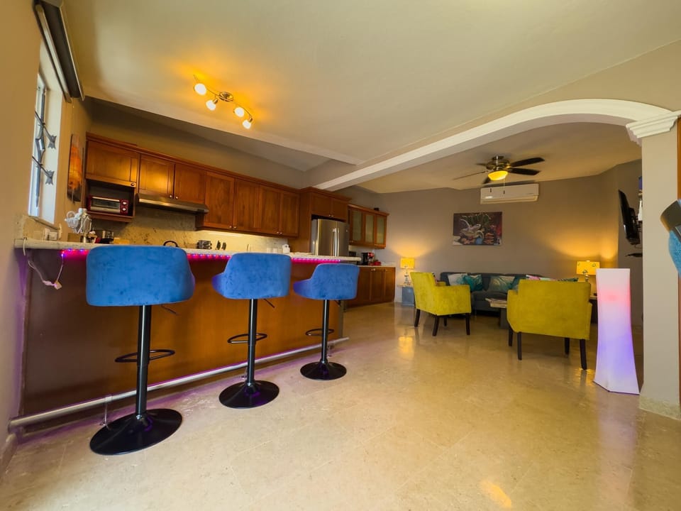 Blue velvet bar stools line the kitchen island with wooden cabinets and marble flooring throughout the living area.