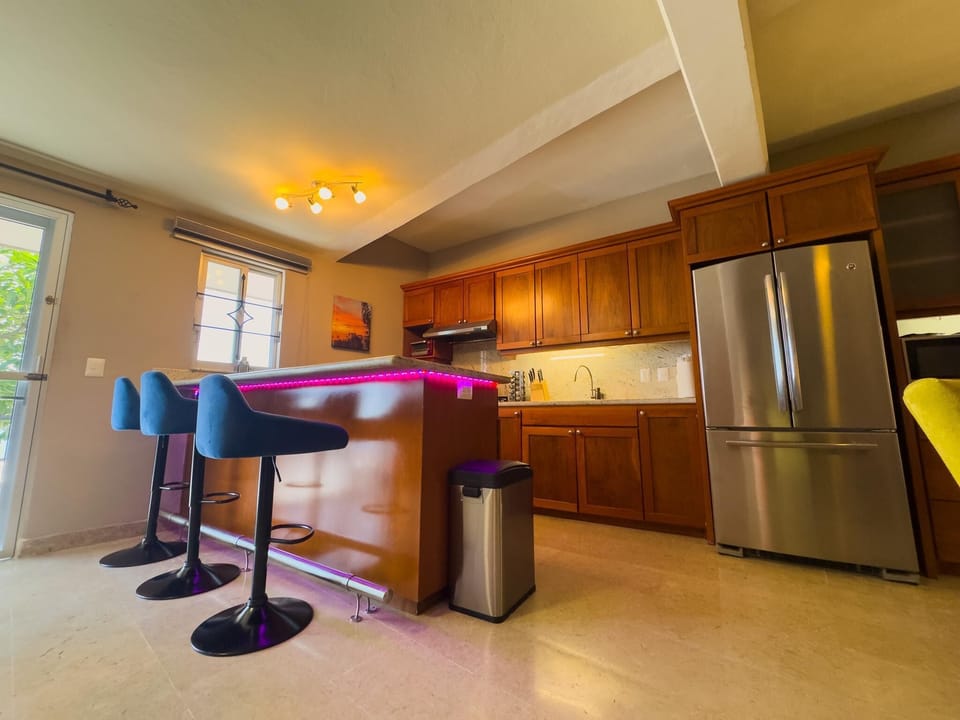 Kitchen island with purple LED lighting, blue swivel bar stools, and warm wood cabinetry against a marble floor.