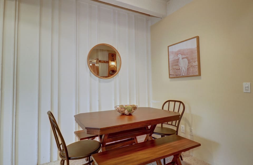 A small dining area with a wooden table, two chairs, and a bench. A framed picture and a round mirror adorn the light-colored walls. A bowl with fruits is placed on the table.