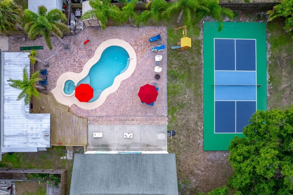 An aerial view of a vibrant backyard featuring a uniquely shaped swimming pool with bright red umbrellas, adjacent to a well-maintained tennis court. The surrounding area is adorned with lush palm trees and ample lounging space, perfect for relaxation and