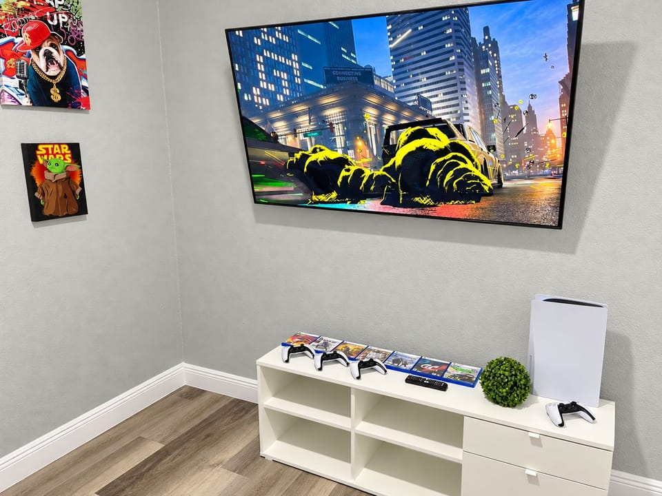Big screen, PS5, ready to play. A wall-mounted Smart TV over a PlayStation 5 and a lineup of games. Plug in and play from the first night, no setup needed.