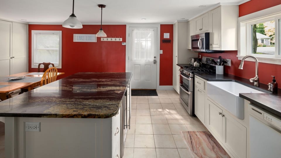 A kitchen with red walls, white cabinets, a large island with a granite countertop, stainless steel appliances, and a farmhouse sink. A door and window provide natural light.