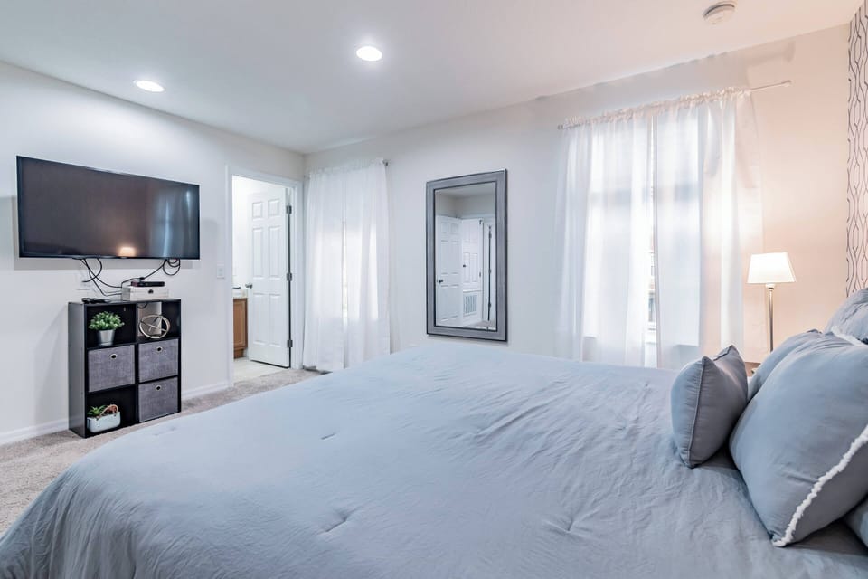 Unwind in this thoughtfully designed bedroom with a plush gray bed and soft pillows, a stylish wall-mounted TV, and natural light streaming through sheer curtains for a serene and calming atmosphere.