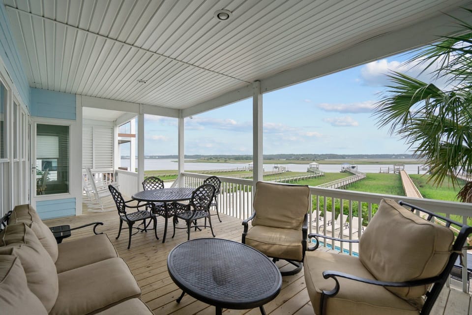 Covered porch with cushioned seating, outdoor dining tables, white railings, marsh and waterway views, pool below, palm tree, and wide coastal scenery.