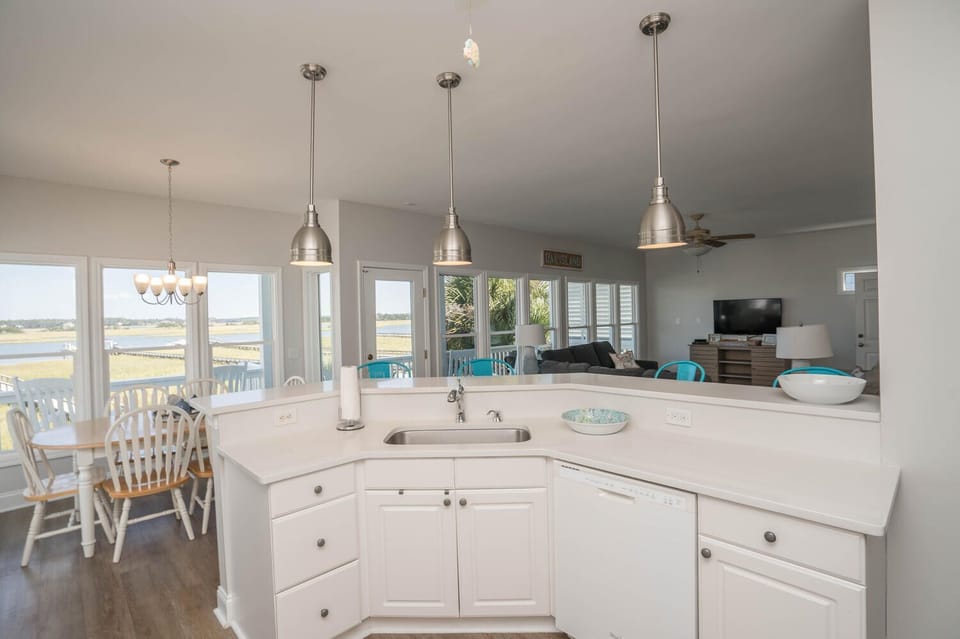 Bright white kitchen with breakfast bar, pendant lights, stainless appliances, ceiling fan, wood-look floors, large windows, and beautiful waterway views.