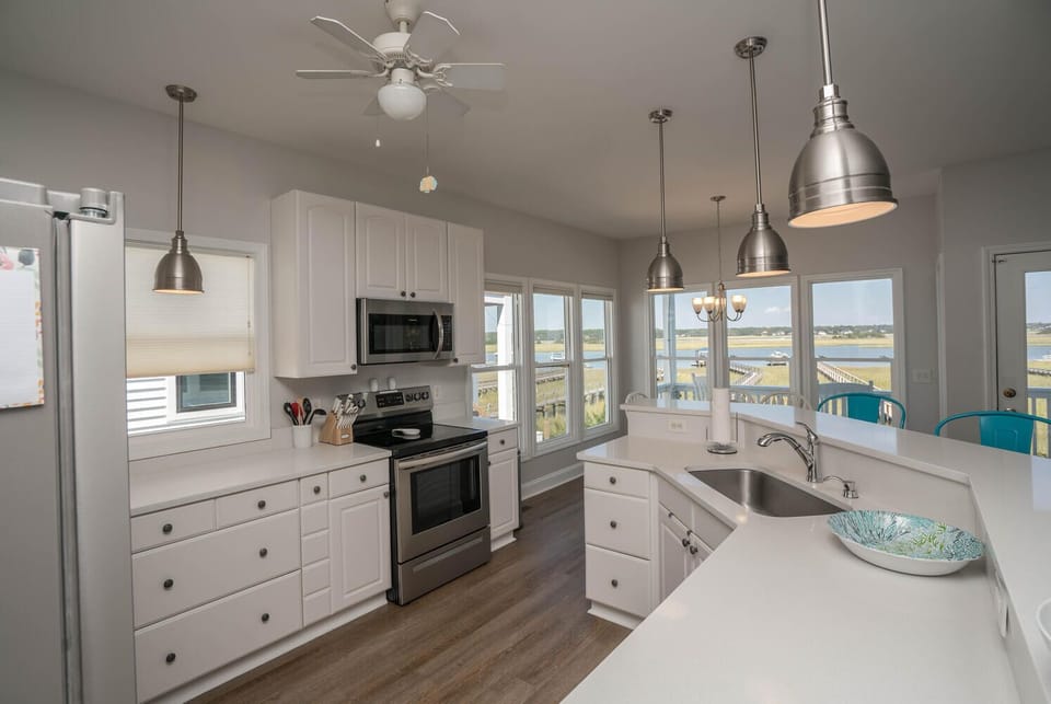 Bright white kitchen with breakfast bar, pendant lights, stainless appliances, ceiling fan, wood-look floors, large windows, and beautiful waterway views.