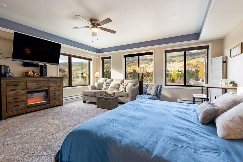 A spacious bedroom with large windows, a fireplace, a wall-mounted TV, a bed with blue bedding, a corner couch, a small desk, and scenic views of a mountainous landscape outside.