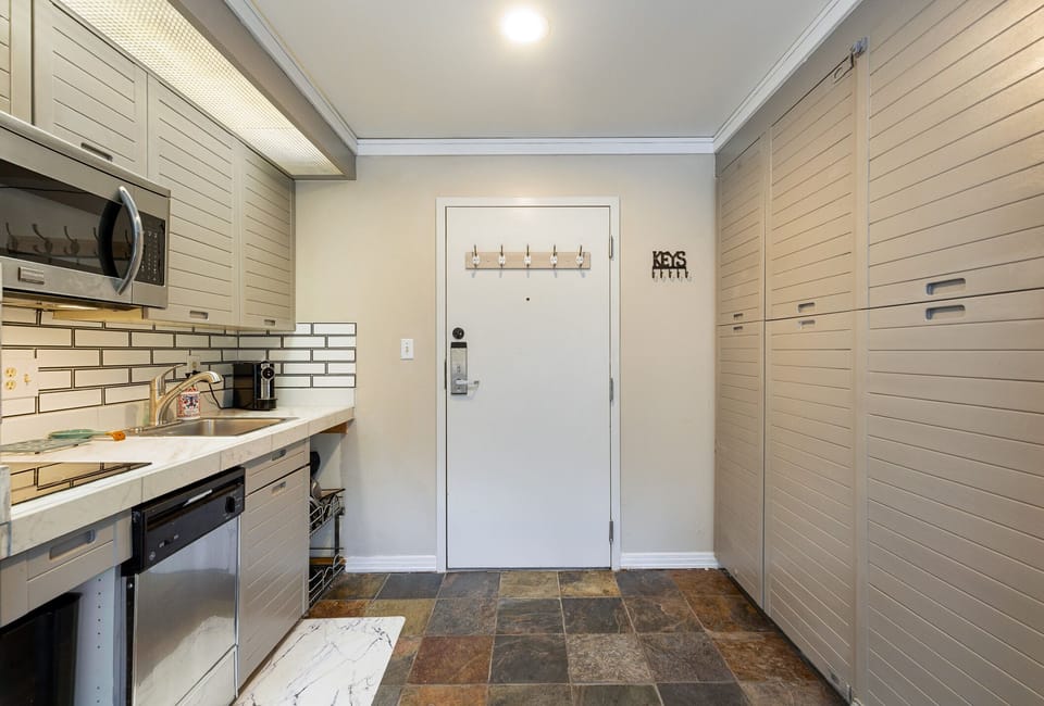 A compact kitchen area with light gray cabinets, a microwave, a dishwasher, a tiled backsplash, and black tiled flooring. The room has a door with hooks for hanging items.