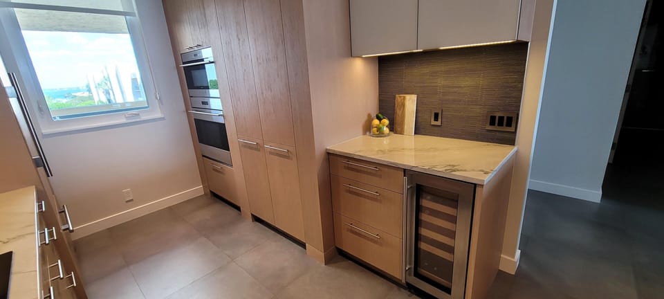 Kitchen corner featuring light wood cabinetry, marble countertop, built-in wine cooler, and textured dark backsplash undercabinet lighting.