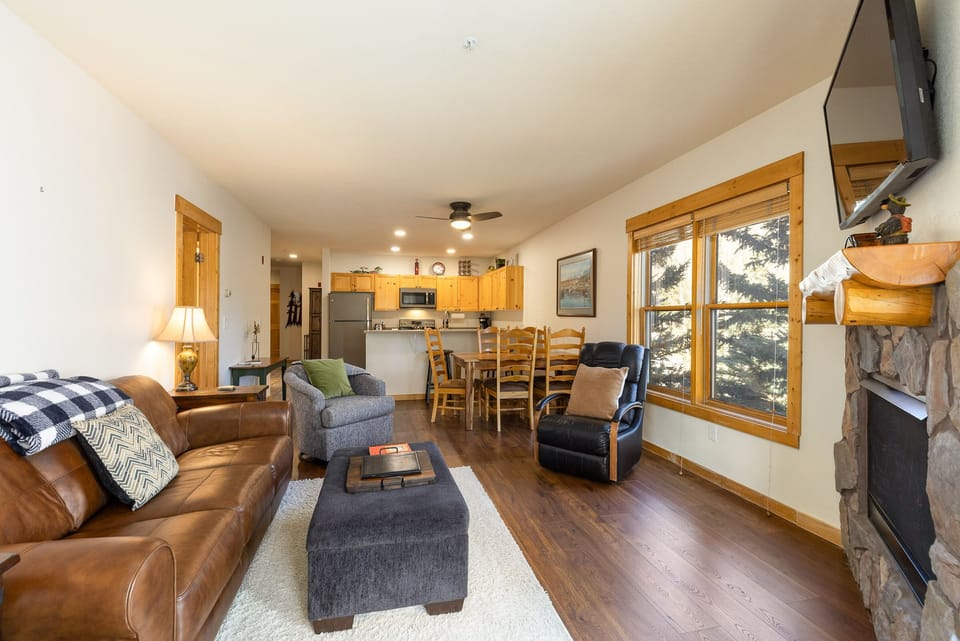 A cozy living room with a leather sofa, armchair, and footrest faces a wall-mounted TV. A dining table and kitchen are visible in the background. Large windows allow natural light to fill the space.