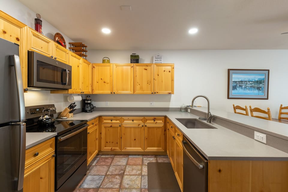 A modern kitchen with wooden cabinets, stainless steel appliances, a microwave, coffee maker, double sink, and a L-shaped counter. The floor has tiled sections and a painting hangs on the wall.