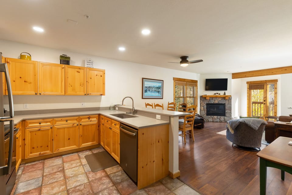 An open-concept kitchen and living room with wooden cabinetry, stainless steel appliances, a stone fireplace, and a wall-mounted TV. The space includes a dining area and leads to a patio through glass doors.