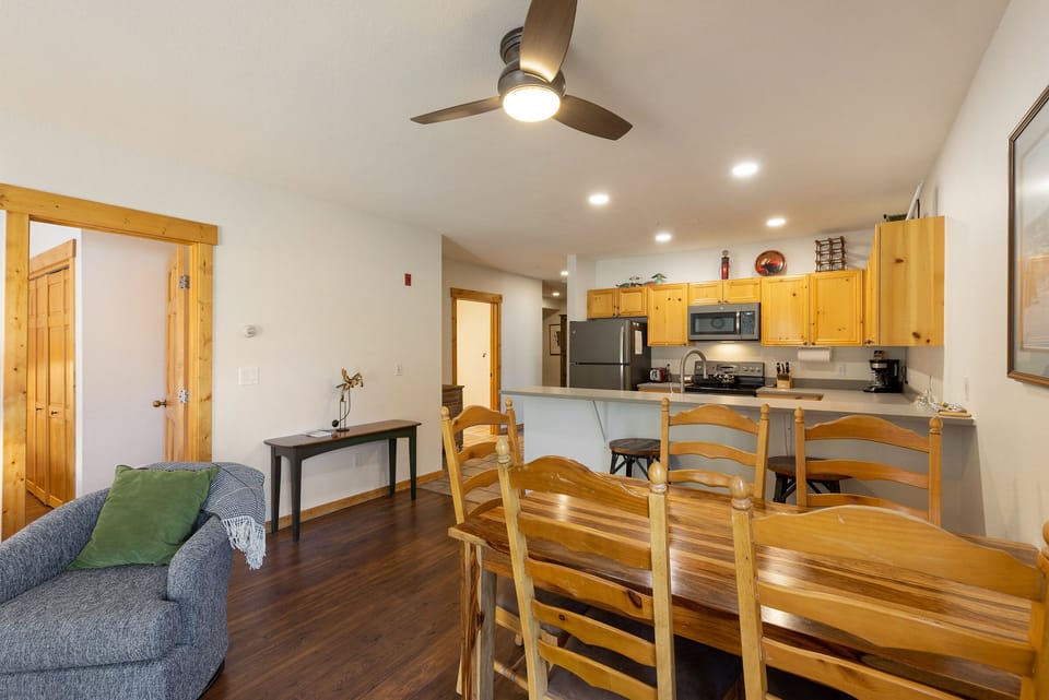 A cozy kitchen and dining area with wooden cabinetry, stainless steel appliances, a ceiling fan, and a wooden dining table with six chairs. An adjacent living area includes a cushioned armchair with a blanket.