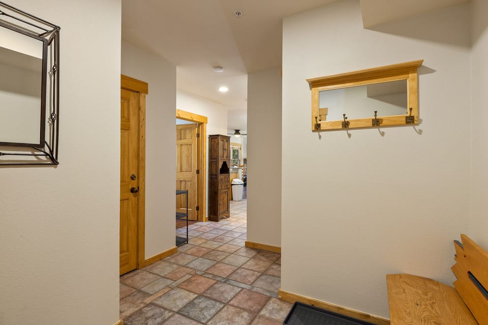 A view of a hallway with tiled flooring, light beige walls, wooden doors, a wall-mounted mirror adjacent to hooks, and a bench on the right. At the end, there is a glimpse of a room with more decor.