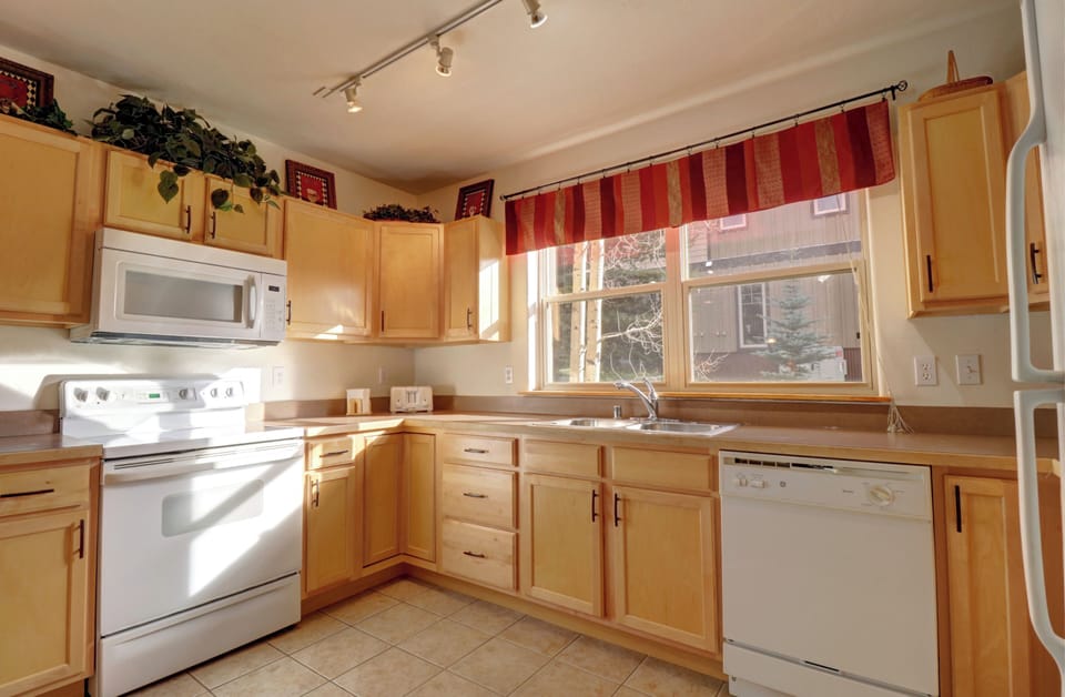 A bright kitchen with wooden cabinets, a white stove, microwave, dishwasher, and a large window featuring striped red curtains. Countertop includes a sink and various small appliances. Floor is tiled.