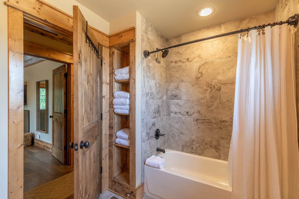 Fresh towels stacked and ready, rustic wood doors, and a stone-tiled tub-shower combo for a relaxing mountain retreat.
