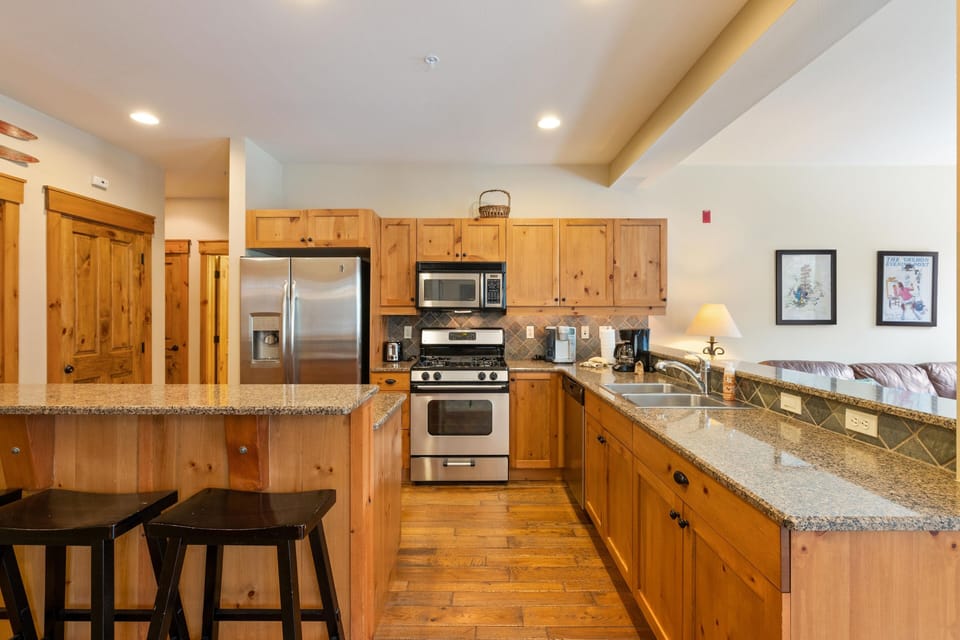 A kitchen features wooden cabinets, stainless steel appliances, a granite countertop with two bar stools, and overhead lighting. Adjacent is a living area with a couch, side table, and framed artworks.