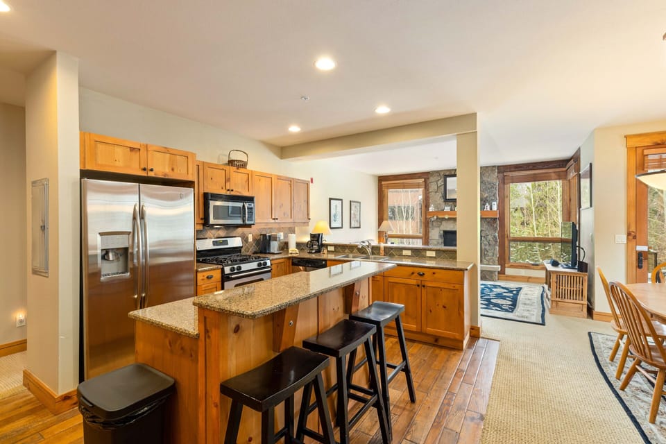 Modern kitchen and living area with wooden cabinetry, granite countertops, stainless steel appliances, and bar stools at the island. The living area includes a large window and stone fireplace.