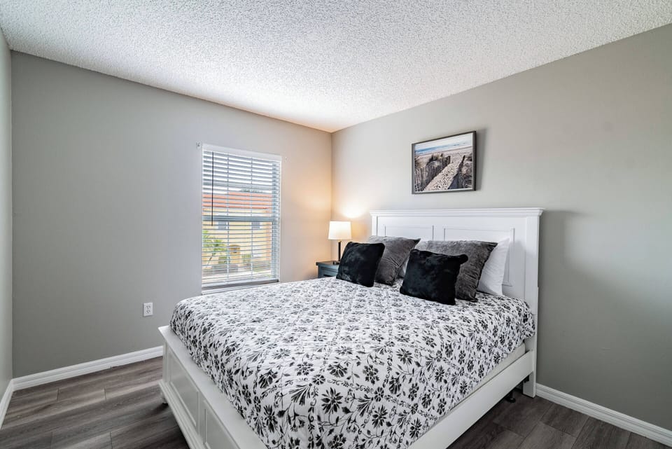 Experience restful nights in this cozy bedroom, featuring a plush white bed adorned with a stylish black-and-white patterned duvet. Natural light streams through the window, enhancing the soft gray walls and inviting ambiance.