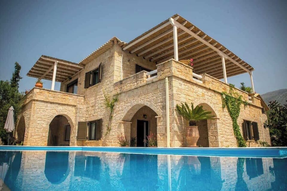 View of the main villa from the pool, showing the stone exterior and both balcony areas overlooking the surrounding landscape.