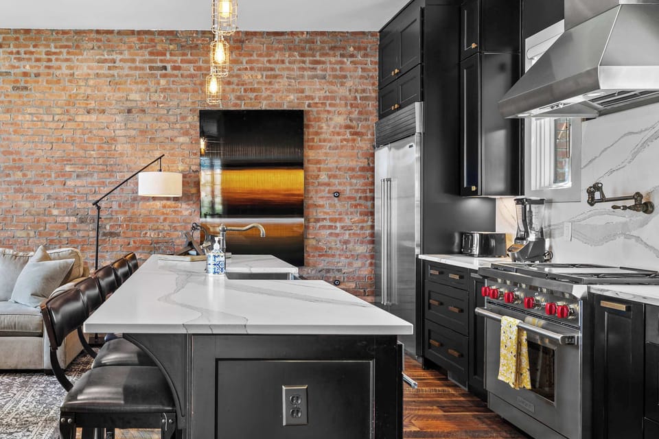 Spacious and inviting living room featuring comfortable sectional seating, exposed brick walls, a Smart TV, and an open-concept layout that flows seamlessly into the kitchen and dining area.

