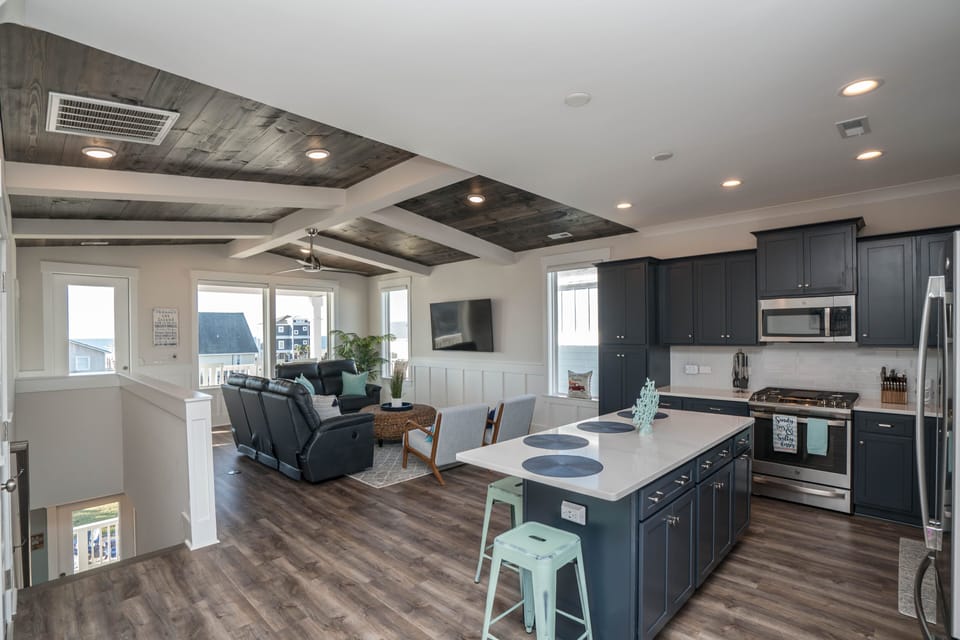 Modern kitchen with navy cabinets, large island, mint stools, stainless appliances, gas range, dining table, chandelier, tall windows, and wood floors.