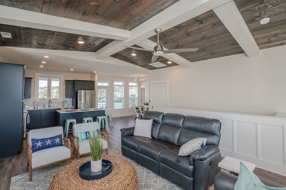 Bright living room with wood-beam ceiling, wall TV, leather seating, accent chairs, round woven coffee table, ceiling fan, large windows, and deck access.