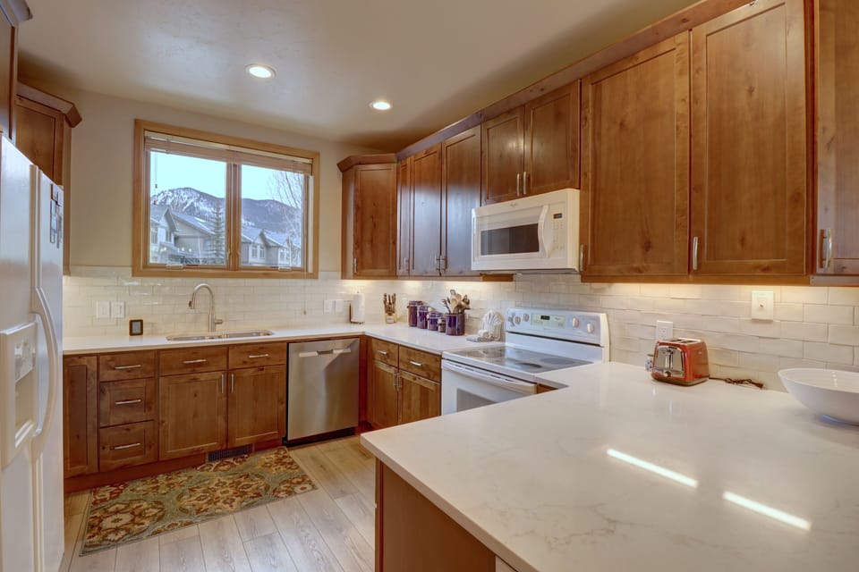 A modern kitchen with wooden cabinets, white countertops, a built-in microwave, a dishwasher, and a window with a mountain view. A toaster and dishware are on the counter.