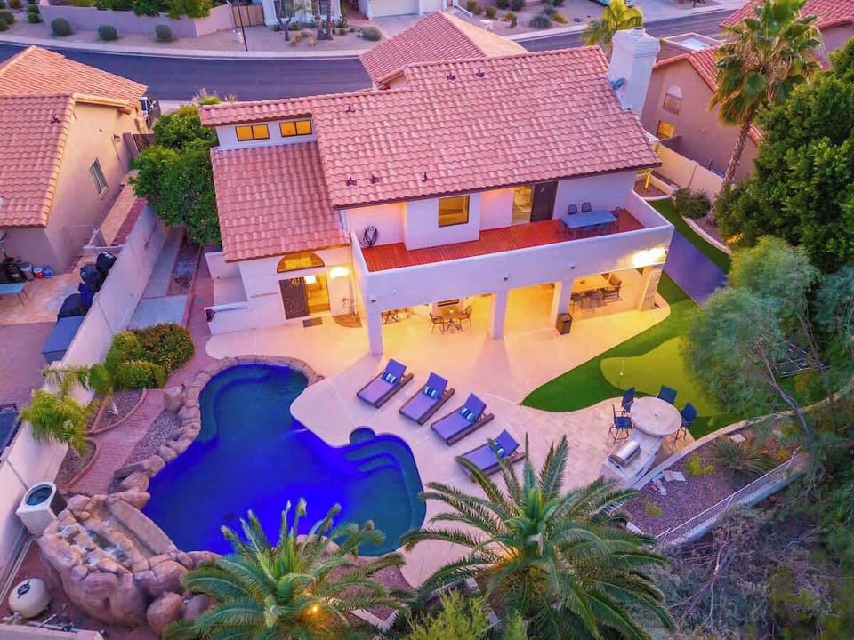 Aerial view of a vacation rental featuring a custom pool with rock waterfall, spacious covered patio with dining area, and landscaped backyard in a quiet residential neighborhood.