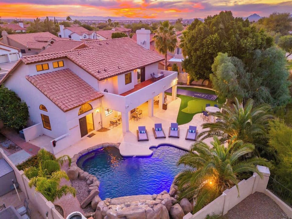 Aerial view of a white stucco rental home with terra cotta tile roof, featuring a resort-style pool, spacious patio with lounge chairs, and lush desert landscaping, set against mountains and a vibrant sunset sky.