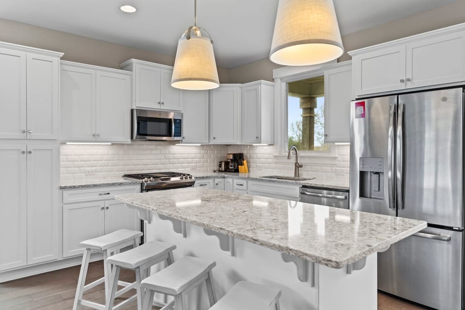 Bright coastal kitchen with white cabinetry, granite countertops, subway tile backsplash, stainless appliances, large island with seating, pendant lighting, and window over the sink.