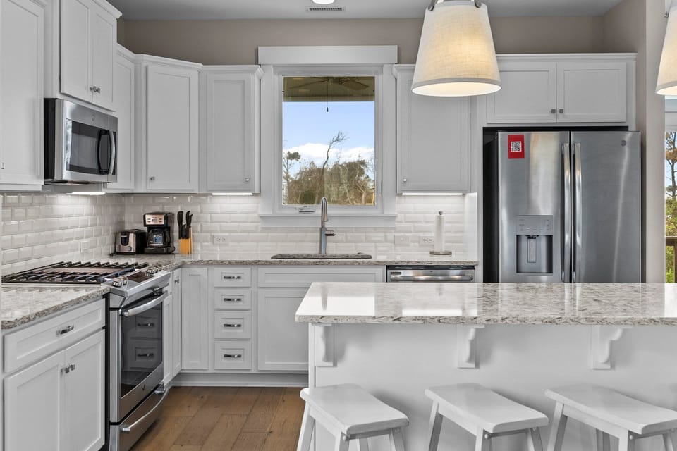 Bright coastal kitchen with white cabinetry, granite countertops, subway tile backsplash, stainless appliances, large island with seating, pendant lighting, and window over the sink.