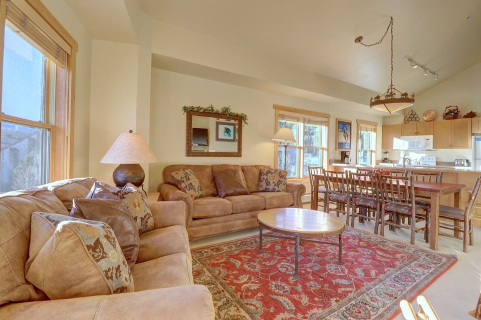 A cozy living room with beige sofas, a round wooden coffee table on a red patterned rug, a dining table with chairs, and a kitchen in the background.