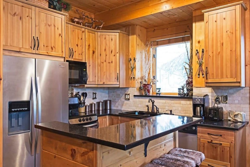 The lovely kitchen area boasts an island with bar seating for two, stainless steel appliances, wood finishes, a tile backsplash and nature views from the kitchen sink.